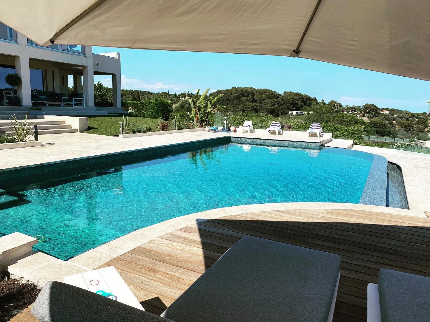 Piscine privée bordée de travertin et terrasse ipé, vue panoramique sur les collines boisées de la Côte d'Azur sous un parasol