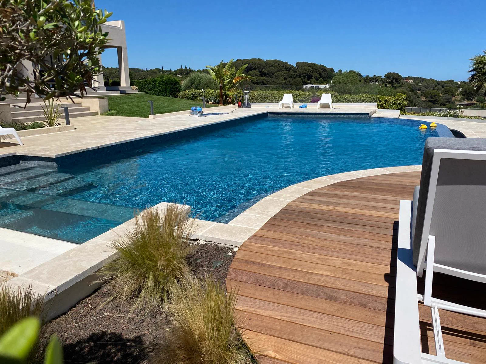 Le même jardin réalisé : piscine bordée de travertin, terrasse en bois courbe et graminées sous un ciel bleu — réalisation Influence Paysage